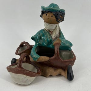 May include: A ceramic figurine of a person wearing a teal hat and matching coat with a white scarf. The figure has dark hair and is positioned behind two brown, basket-shaped containers with white interiors, all crafted from glazed clay.