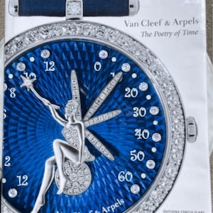 May include: A hardcover book titled Van Cleef & Arpels The Poetry of Time featuring a large watch face on the cover. The watch has a deep blue guilloché dial, a diamond-encrusted bezel, and a silver fairy figure with a star-tipped wand as a hand.