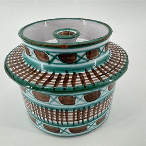 May include: A ceramic butter crock with a white base, featuring hand-painted teal green lines and brown rectangular accents. The vessel has a wide, flared rim and a central lid with a small knob handle, decorated with a repeating geometric pattern.