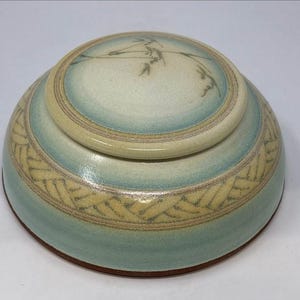 May include: A round ceramic lidded jar with a smooth, glossy finish. The piece features a soft gradient of pale blue and cream tones, accented by a textured, woven-style pattern around the base and a delicate, hand-painted botanical motif on the lid.