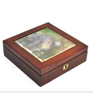 May include: A square wooden box with a dark brown finish and a hinged lid. The center of the lid features an inset square tile with an abstract, multi-colored pattern of gray, green, yellow, and orange. A small brass keyhole is centered on the front.