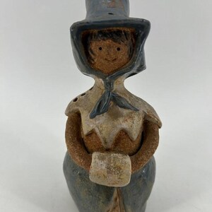 May include: A ceramic figurine of a person wearing a blue bonnet and a matching blue skirt with a textured brown bodice. The figure has a light brown face with simple facial features and holds a small, rectangular, light-colored object in front.