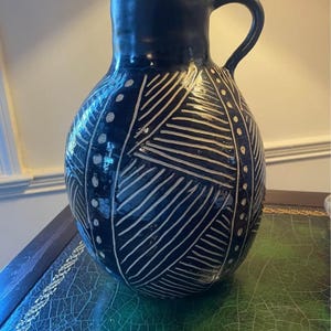 May include: A dark navy blue ceramic pitcher with a rounded body and a single handle. The surface features hand-carved white geometric patterns consisting of diagonal lines and vertical columns of dots that wrap around the entire vessel.