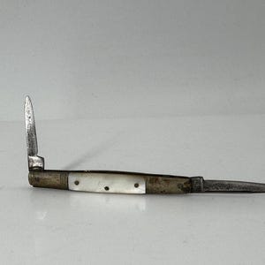 May include: A folding pocket knife with two metal blades extended at opposite ends. The handle features a central inlay of iridescent white mother-of-pearl material secured by three small metal pins, flanked by aged, tarnished brass-toned metal bolsters.