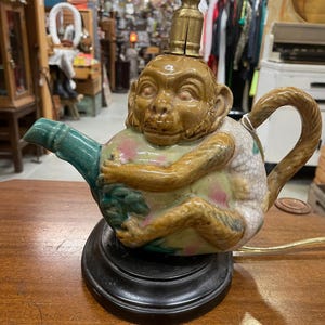 May include: A whimsical lamp featuring a ceramic teapot base with a monkey figure. The teapot is painted with pastel colors and has a green spout and handle. The lamp sits on a black base and has a brass lamp fixture.
