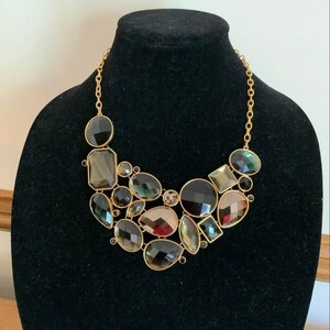 May include: A statement necklace featuring a gold-tone chain and a cluster of faceted, multi-shaped stones in shades of black, charcoal, and deep brown. The stones are arranged in a layered, asymmetrical bib design with a polished finish.