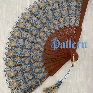 Può includere: Un ventaglio pieghevole con stecche in legno marrone e una foglia in pizzo blu e oro con motivo floreale. Il ventaglio include un anello dorato alla base con una nappa blu e oro. Il testo sull'immagine recita Ventaglio blu e oro, Pattern e (c) Edda Guastalla 2026.
