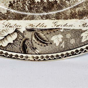 May include: A vintage brown and white ceramic plate with a black and white image of the Washington Statue in Boston's Public Garden. The plate is decorated with a floral border and the text "Washington Statue. Public Garden. Boston. J. Me D & S."