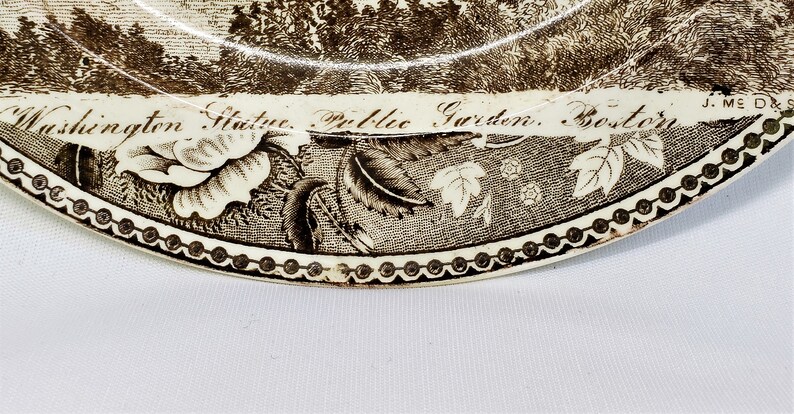 May include: A vintage brown and white ceramic plate with a black and white image of the Washington Statue in Boston's Public Garden. The plate is decorated with a floral border and the text "Washington Statue. Public Garden. Boston. J. Me D & S."