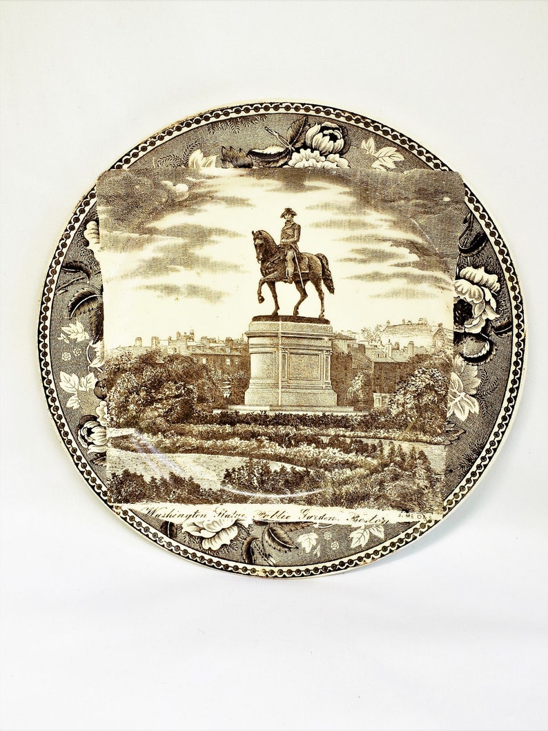 May include: A brown and white ceramic plate with a black and white image of a statue of George Washington on horseback in Washington Square Park, New York City. The plate has a decorative border with floral designs. The text "Washington Square Public Garden, New York" is printed on the bottom of the plate.