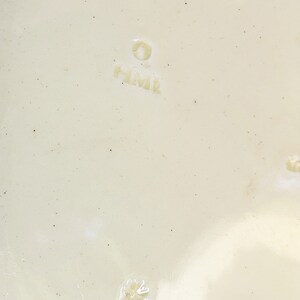 May include: Close-up of a white ceramic plate with a small, white flower design and the text "H.M.K." in a circle.