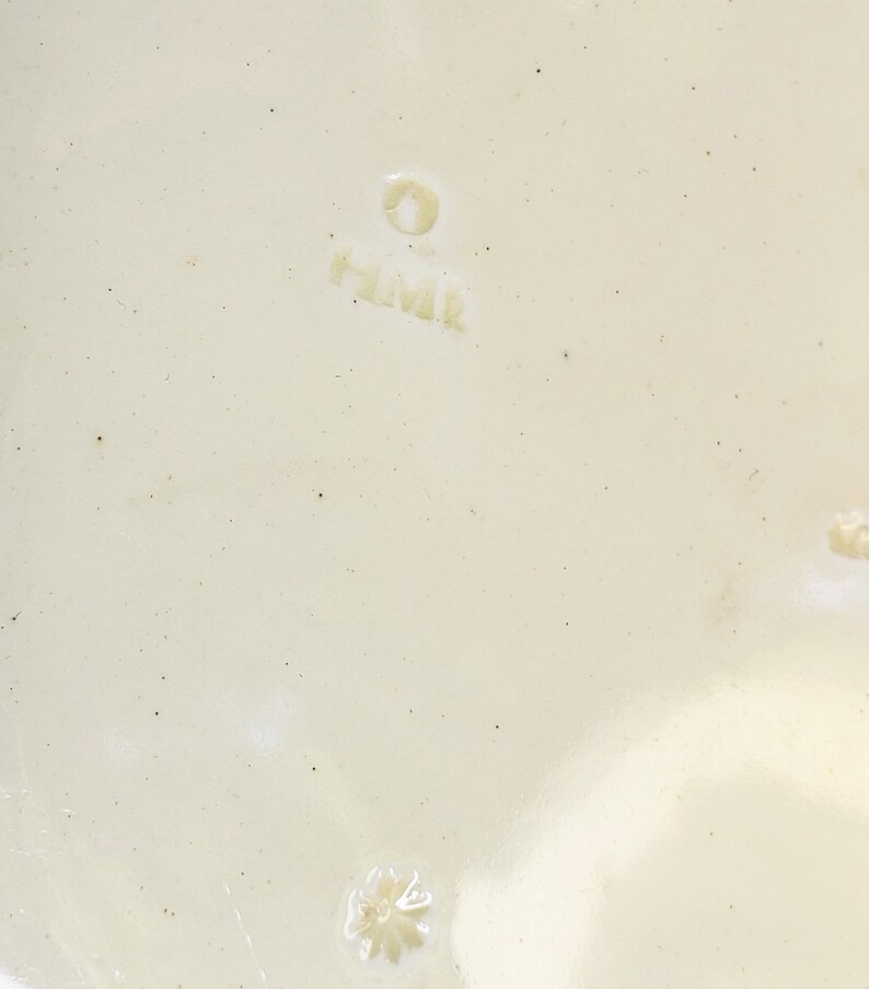 May include: Close-up of a white ceramic plate with a small, white flower design and the text "H.M.K." in a circle.