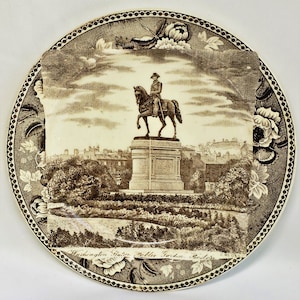 May include: A brown and white ceramic plate with a black and white image of the Washington Monument in Boston's Public Garden. The plate has a decorative border with floral designs.