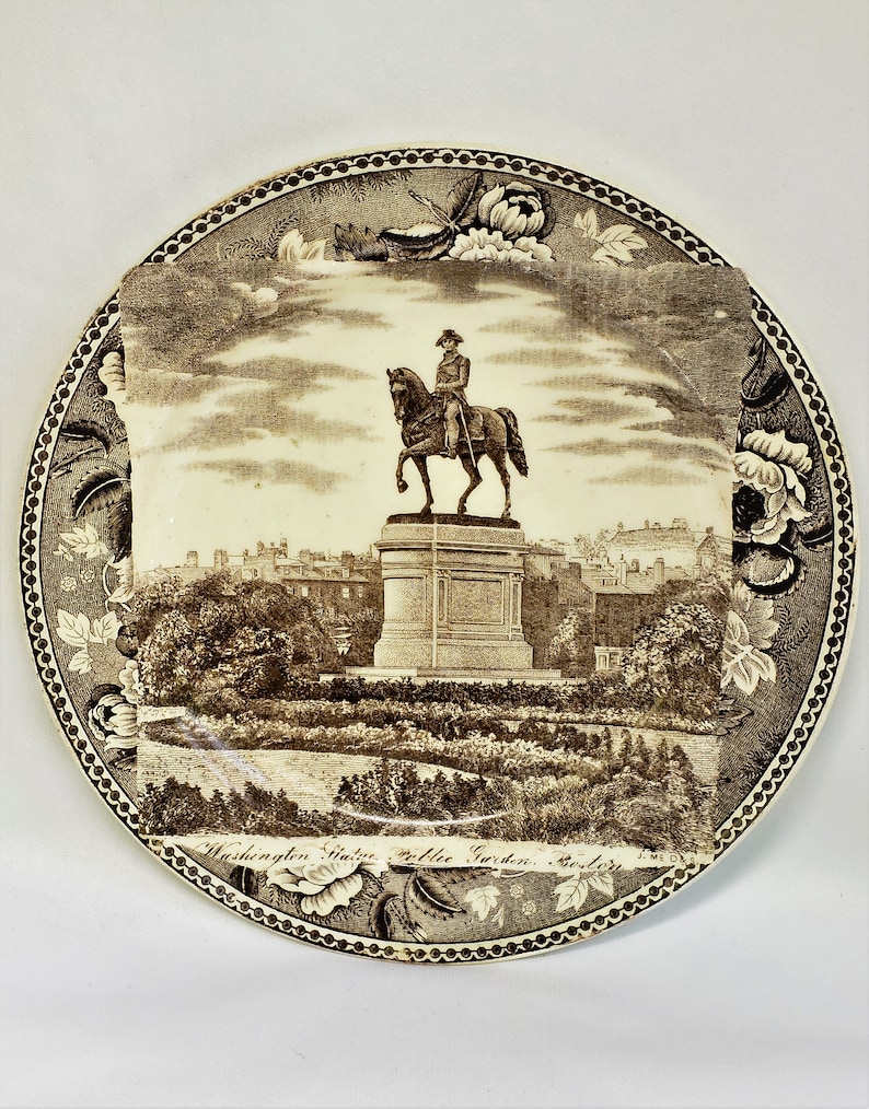 May include: A brown and white ceramic plate with a black and white image of the Washington Monument in Boston's Public Garden. The plate has a decorative border with floral designs.
