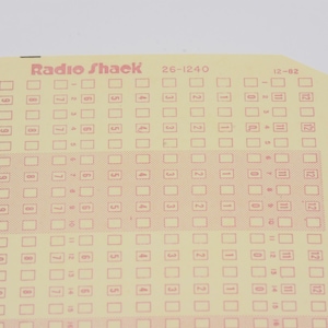 May include: A cream-colored Radio Shack 26-1240 card featuring a grid of numbered boxes and columns. The card includes a vertical row of dark brown rectangular marks along the left edge and a central horizontal band with a light orange grid pattern.