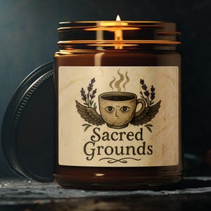 Peut inclure: Une bougie allumée dans un bocal en verre brun avec un couvercle noir. L'étiquette présente une tasse à café avec des ailes et les mots "Sacred Grounds". La bougie est sur une surface sombre avec un fond sombre.
