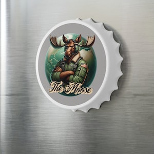 Puede incluir: Un imán blanco con forma de tapón de botella con un personaje de dibujos animados de alce. El alce lleva una chaqueta verde y tiene los brazos cruzados. El fondo es un globo terráqueo. El texto "The Moose" está debajo.