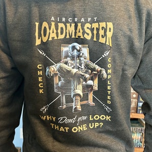 Pode incluir: Um moletom com capuz cinza-carvão com um gráfico nas costas de um piloto com traje de voo e capacete sentado em uma cadeira. O texto diz AIRCRAFT LOADMASTER, CHECK COMPLETED e WHY DONT YOU LOOK THAT ONE UP? com duas flechas cruzadas.
