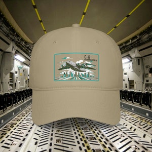 May include: A beige baseball cap with a patch featuring a C-17 Globemaster III airplane flying over a city skyline. The patch has a teal border and the text "C-17 Globemaster III" in black.