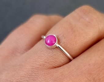Könnte beinhalten: Ein Silberring mit einem kleinen, runden, leuchtend pinkfarbenen Cabochon-Stein, der in einer einfachen Metallfassung sitzt. Der Ring ist dünn und poliert und weist eine glatte, metallische Oberfläche auf, die den Finger umschließt.