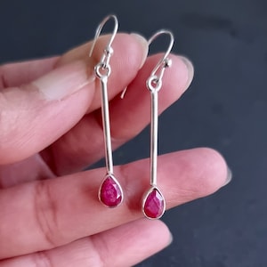 Könnte beinhalten: Ein Paar silberner Ohrhänger. Jeder Ohrring hat einen langen, schlanken Silberstab mit einem tropfenförmigen, facettierten, leuchtend roten Edelstein am unteren Ende. Die Ohrringe werden von einem silbernen Haken gehalten.