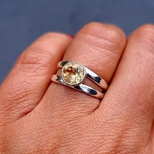 Peut inclure: Une bague argentée ornée d'une pierre précieuse ronde, facettée et jaune pâle, sertie dans une monture lisse. L'anneau est divisé en deux tiges polies distinctes qui convergent à la base, créant un design structurel simple et épuré.