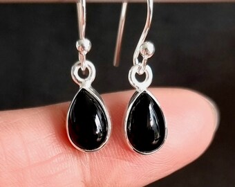 May include: A pair of silver drop earrings with teardrop-shaped, glossy black stones. The earrings have a simple, elegant design, with a small silver ball at the top of the hook. The black stones are set in a silver bezel.