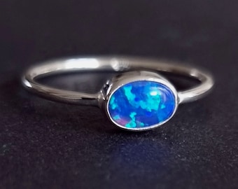 Pode incluir: Um anel de prata com uma gema de opala azul. A opala tem um brilho iridescente azul, verde e roxo.