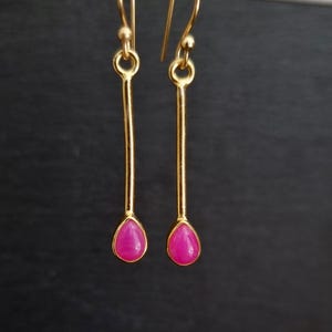 Peut inclure: Paire de boucles d'oreilles pendantes dorées avec une pierre précieuse rose vif en forme de goutte d'eau. Le design est simple et élégant, avec un crochet pour les oreilles. Le fond est sombre et uni.