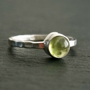 May include: A silver ring featuring a hammered band and a round, light green gemstone. The gemstone is set in a smooth bezel. The ring is shown against a dark backdrop. The ring is a piece of jewellery.