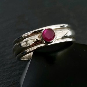 Peut inclure: Une bague en argent avec une pierre précieuse en rubis rouge sertie au centre. La bague a une bande texturée avec trois rainures.