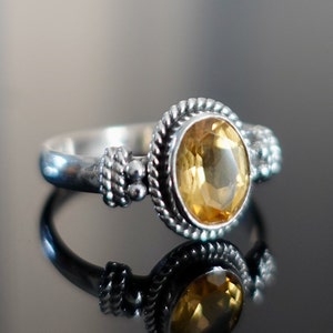 May include: A silver ring with a yellow gemstone set in a detailed bezel. The ring has a rope-like design on the band and small beads on the side.