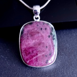 May include: A rectangular pendant featuring a polished, deep magenta and dark green stone with black inclusions, set in a smooth silver frame. The pendant hangs from a silver chain, showcasing a natural, mottled texture across the stone's surface.