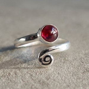 May include: A silver ring with a red gemstone set in a bezel setting. The ring is designed with a swirling band.