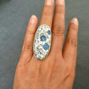 May include: An oval-shaped ring featuring a large, speckled granite-like stone with distinct blue circular inclusions. The stone is set in a simple, polished silver-toned metal bezel that follows the elongated oval contour of the gemstone.