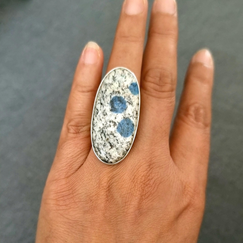May include: An oval-shaped ring featuring a large, speckled granite-like stone with distinct blue circular inclusions. The stone is set in a simple, polished silver-toned metal bezel that follows the elongated oval contour of the gemstone.