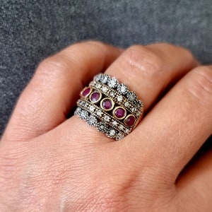 May include: A wide gold ring with a band of alternating silver and gold. The ring is set with a row of seven small, round, dark pink stones.