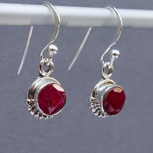 May include: A pair of silver dangle earrings with a red gemstone in the centre of each earring. The gemstones are set in a silver bezel and surrounded by a silver beaded border.