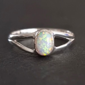 May include: A silver ring featuring a central oval-shaped opal gemstone with iridescent flashes of green, yellow, and orange. The band is polished and splits into two separate arms on each side of the stone, creating an open, bypass-style design.