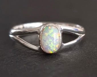 Könnte beinhalten: Ein Silberring mit einem zentralen, ovalen Opal, der irisierende grüne, gelbe und orangefarbene Lichtreflexe aufweist. Die Ringschiene ist poliert und teilt sich zu beiden Seiten des Steins in zwei Arme, was ein offenes Design erzeugt.