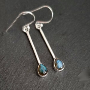 May include: A pair of silver drop earrings featuring teardrop-shaped, iridescent blue and green stones. The earrings have a simple design with a long, slender bar and a hook closure. The stones are set in a silver bezel.