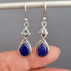 May include: A pair of silver drop earrings featuring a faceted, deep blue gemstone teardrop shape, topped with a small, round, light blue stone. The earrings are suspended from a silver hook.