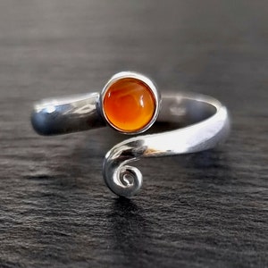 May include: A silver-toned ring with a vibrant orange gemstone. The ring features a swirling design that wraps around the finger, with a round, polished stone set at the top. The ring's design is simple yet elegant.