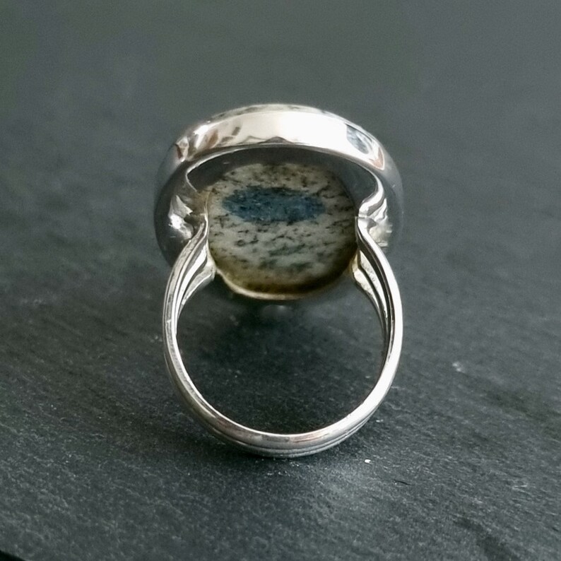 May include: A silver ring featuring a polished, oval-shaped stone with a speckled gray and white pattern and a prominent dark blue, elongated inclusion in the center. The band is constructed from three thin, parallel silver wires.