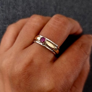 May include: A silver and gold ring with a single red gemstone set in the centre. The ring is worn on a person's finger.