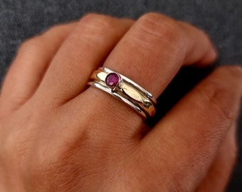 May include: A silver and gold ring with a single red gemstone set in the center. The ring is worn on a person's finger.
