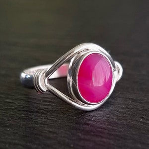 Könnte beinhalten: Ein silberner Ring mit einem leuchtend pinkfarbenen ovalen Edelstein, der in einem gedrehten Drahtdesign gefasst ist.
