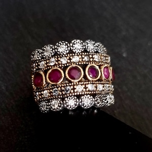 May include: A wide silver and gold ring with multiple bands of small clear stones and a row of larger red stones.