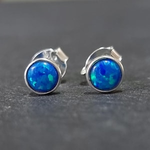May include: A pair of round stud earrings with a vibrant blue opal stone set in a silver bezel. The opal has a mix of blue and green hues. The earrings are displayed on a dark gray surface.
