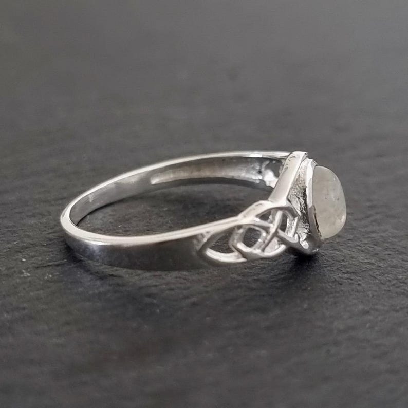 May include: A silver ring with a Celtic knot design on the band and a light-colored, oval-shaped stone. The ring is set against a dark background, highlighting its intricate details and polished finish.
